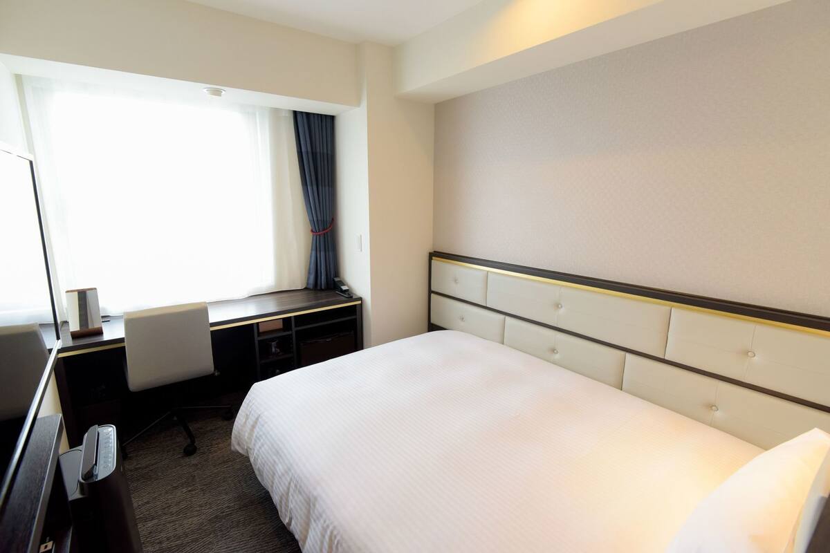 A hotel room with a bed, a desk, a chair, a telephone, and a window with curtains.