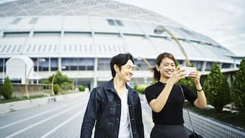 Dos personas tomándose un selfie frente a un estadio moderno con techo de vidrio.
