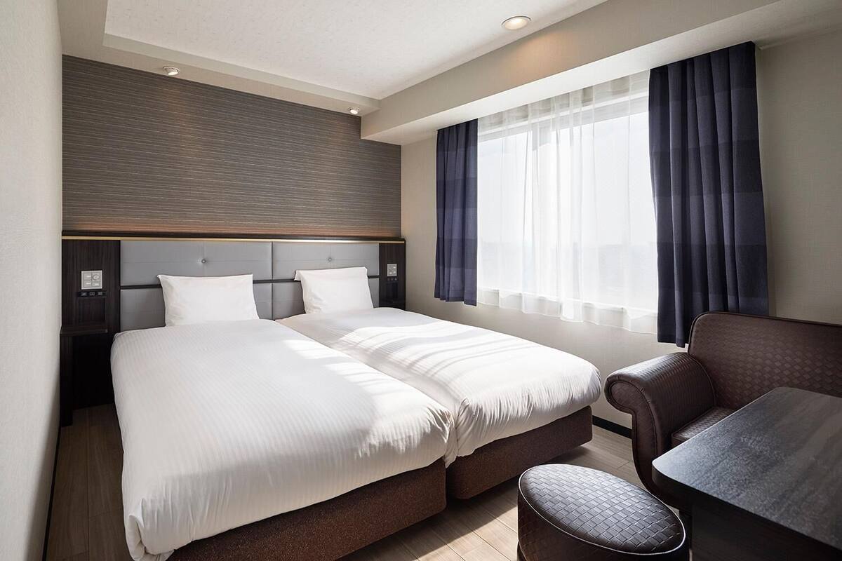 A hotel room with a large bed, a seating area with a chair and ottoman, a control panel, and a window with curtains.