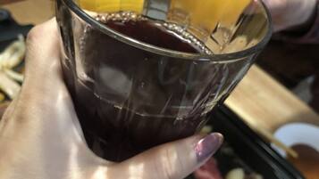 A person holding a glass with a dark liquid, possibly juice, and a tray with various meats and vegetables.