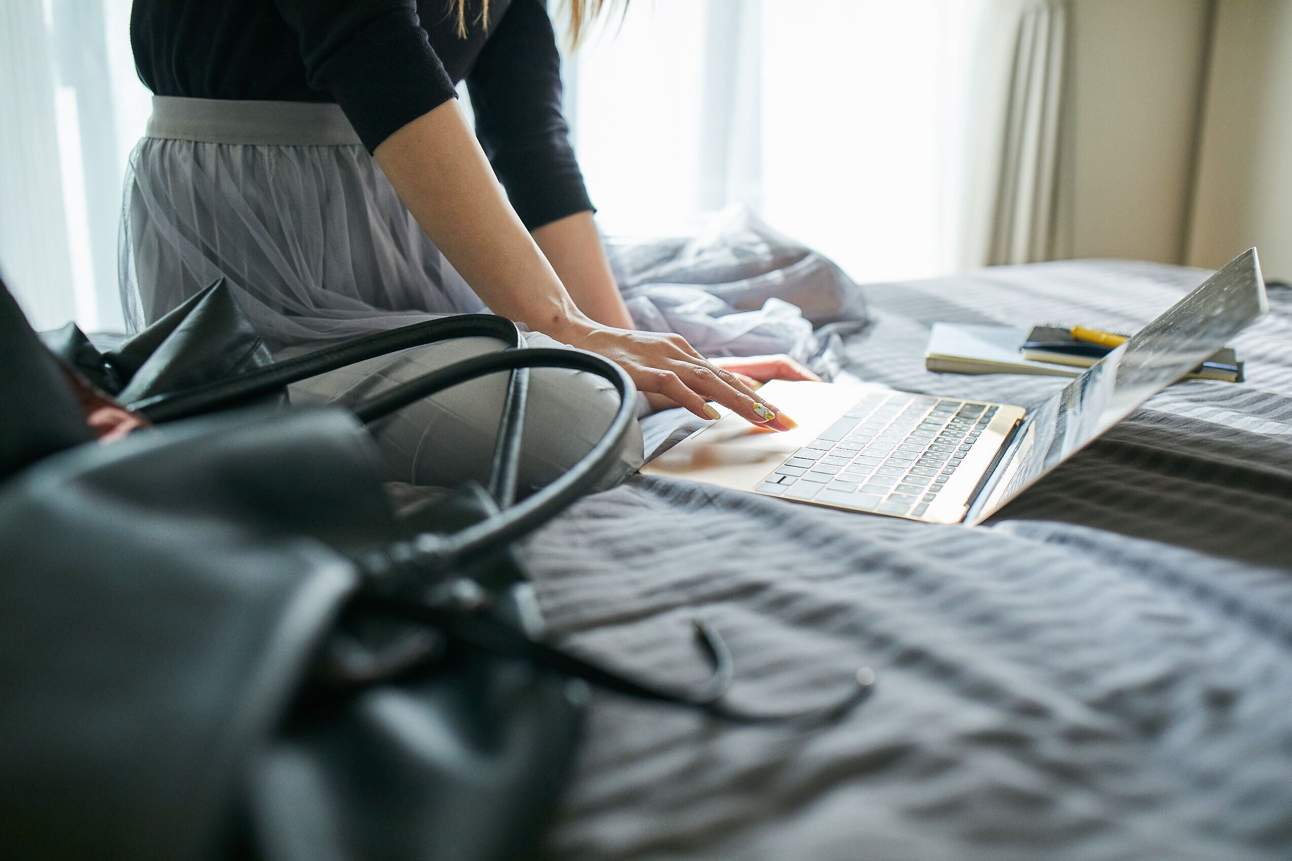 Una persona usando una computadora portátil en una cama, con un bolso negro y una libreta cerca.