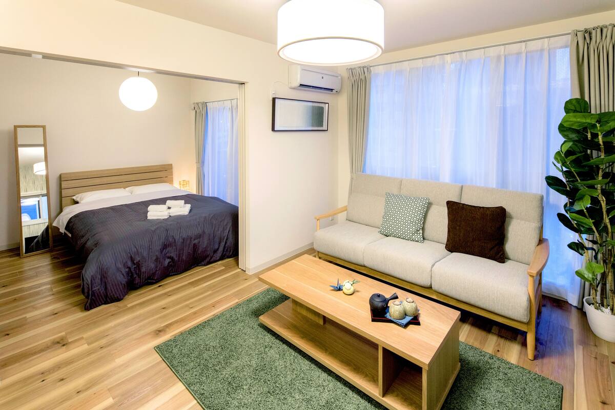 Una habitación de hotel moderna con suelo de madera, cama con ropa de cama oscura, sofá con cojines estampados, una mesa de centro con objetos decorativos y un ventanal con cortinas de gasa.
