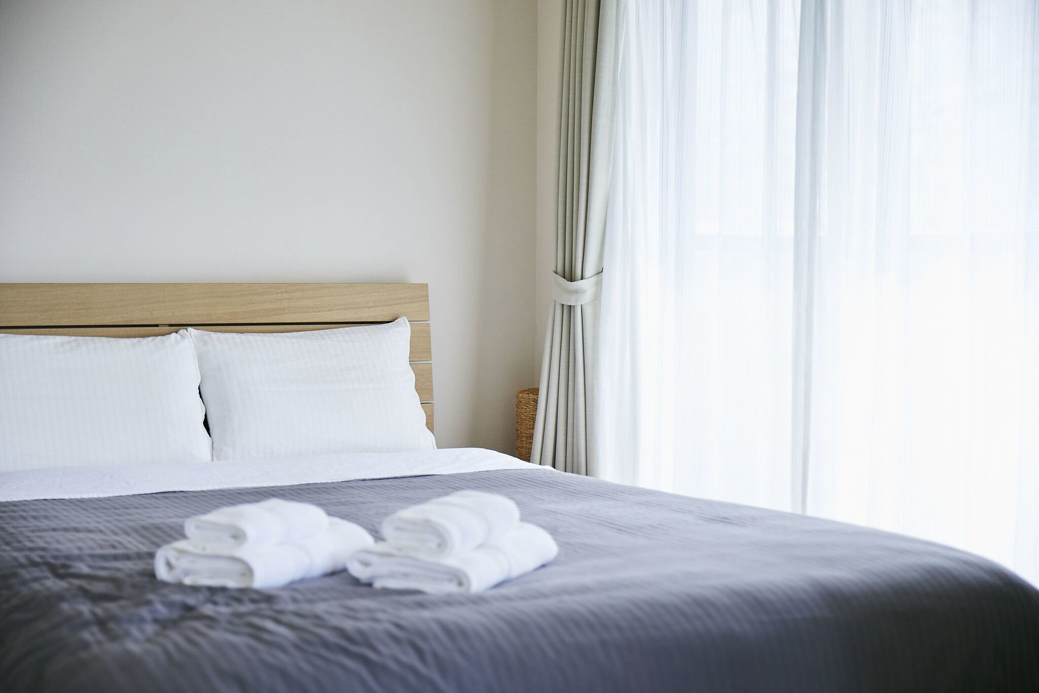 Una cama bien hecha con almohadas blancas y toallas dobladas, un cabecero de madera y cortinas translúcidas que dejan entrar luz natural.