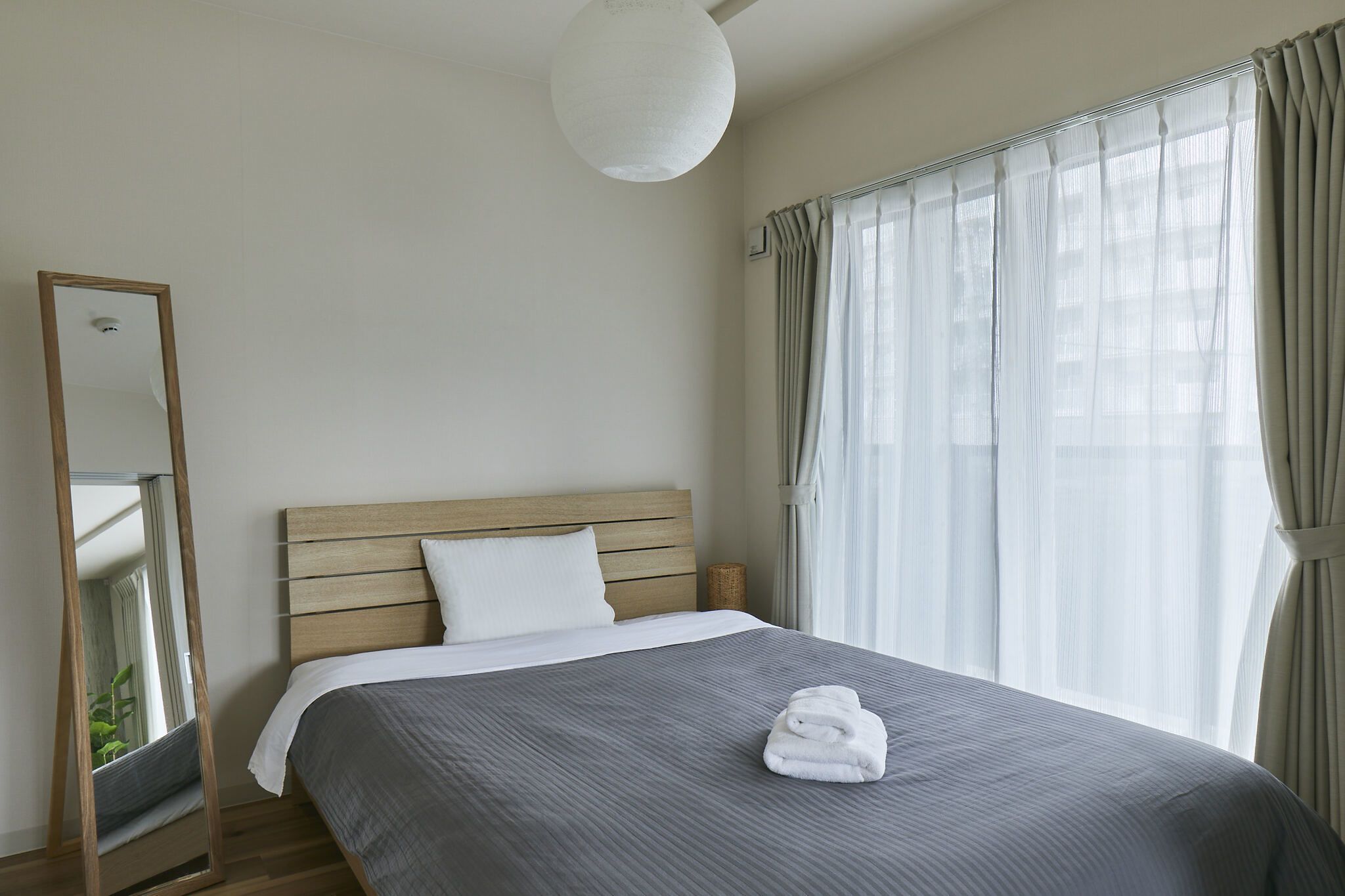 A neatly made bed with a grey bedspread and a folded white towel, a wooden headboard, a large mirror, and sheer curtains on a window.