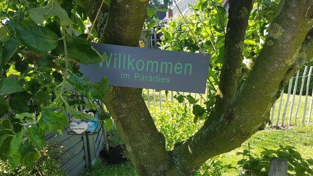 A welcome sign in a garden area with a tree and a metal fence in the background. - Cozy apartment with maritime charm (Langenhorn)