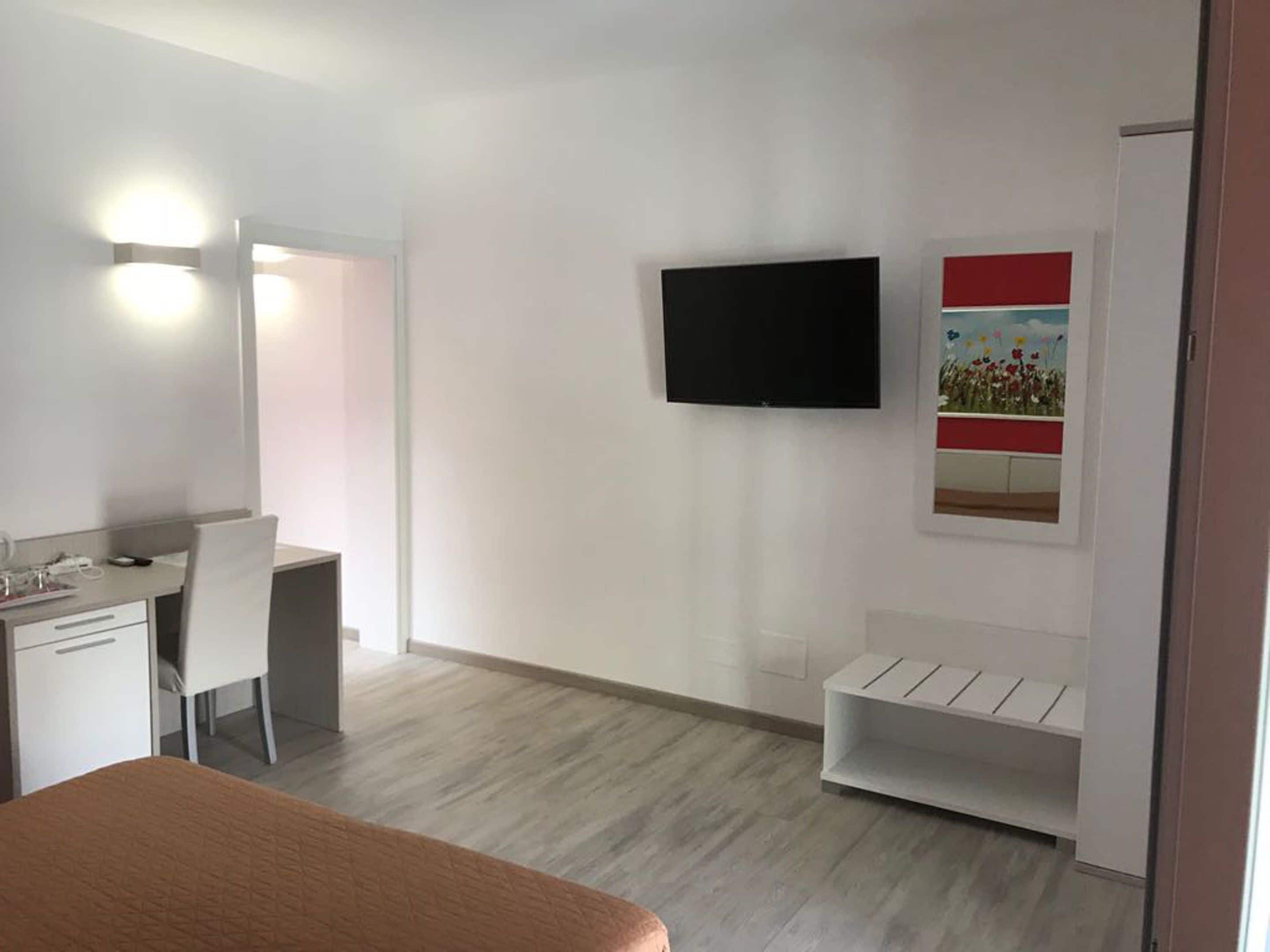 Quarto de hotel moderno com cama, escrivaninha, cadeira, TV fixada na parede e uma pequena prateleira.