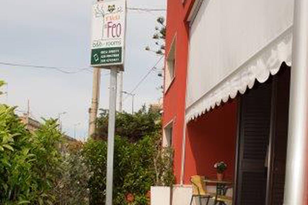 Un estrecho camino empedrado que conduce a un edificio rojo con toldo blanco. Se ve un cartel a la izquierda, y hay sillas y una mesa al aire libre a la derecha.