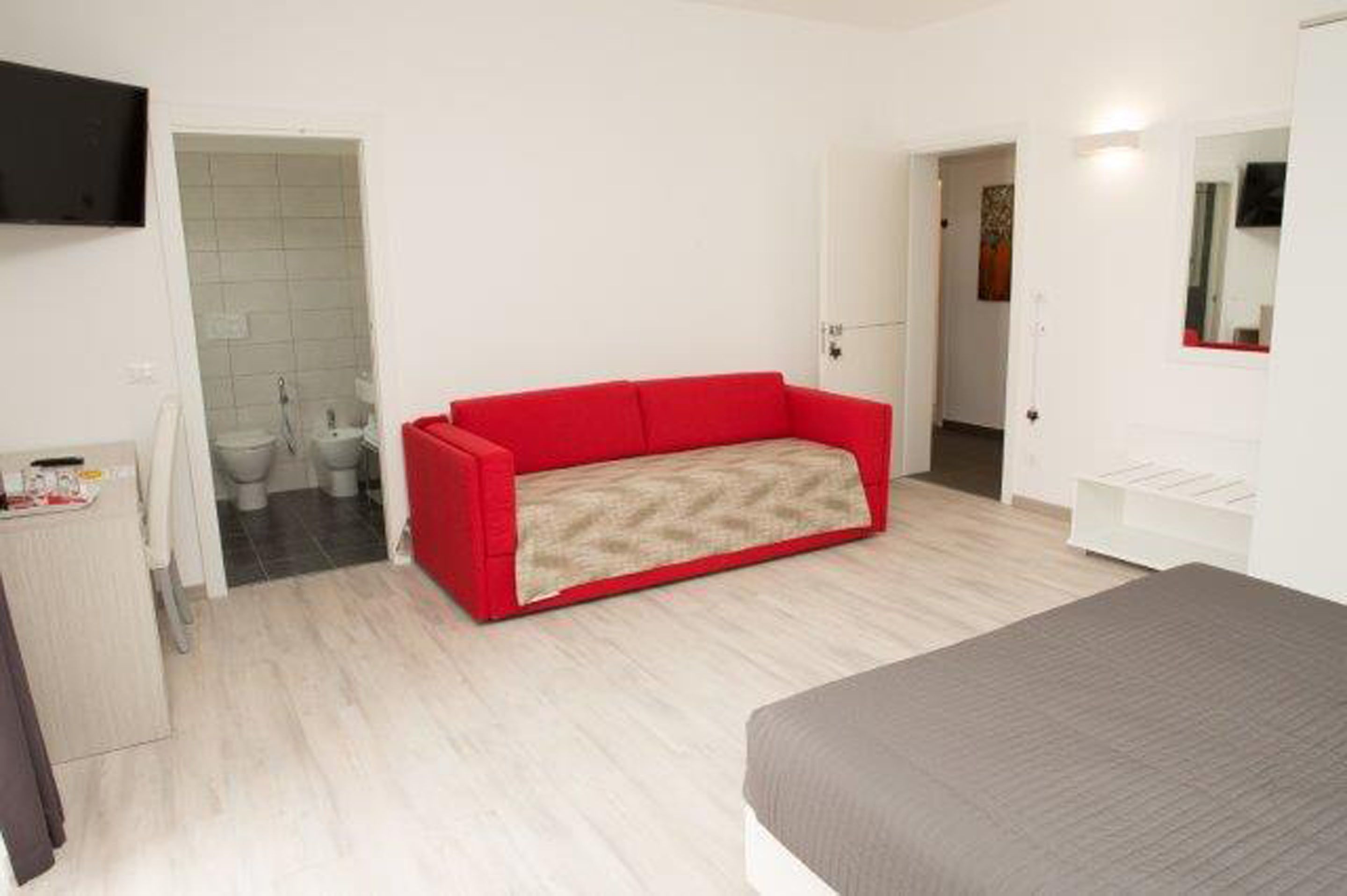 A bedroom with a red sofa, a bed, a TV mounted on the wall, and a bathroom visible through an open door.