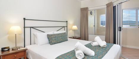 A hotel room with a bed, two bedside lamps, a nightstand, a window with curtains, and a view of the sea.