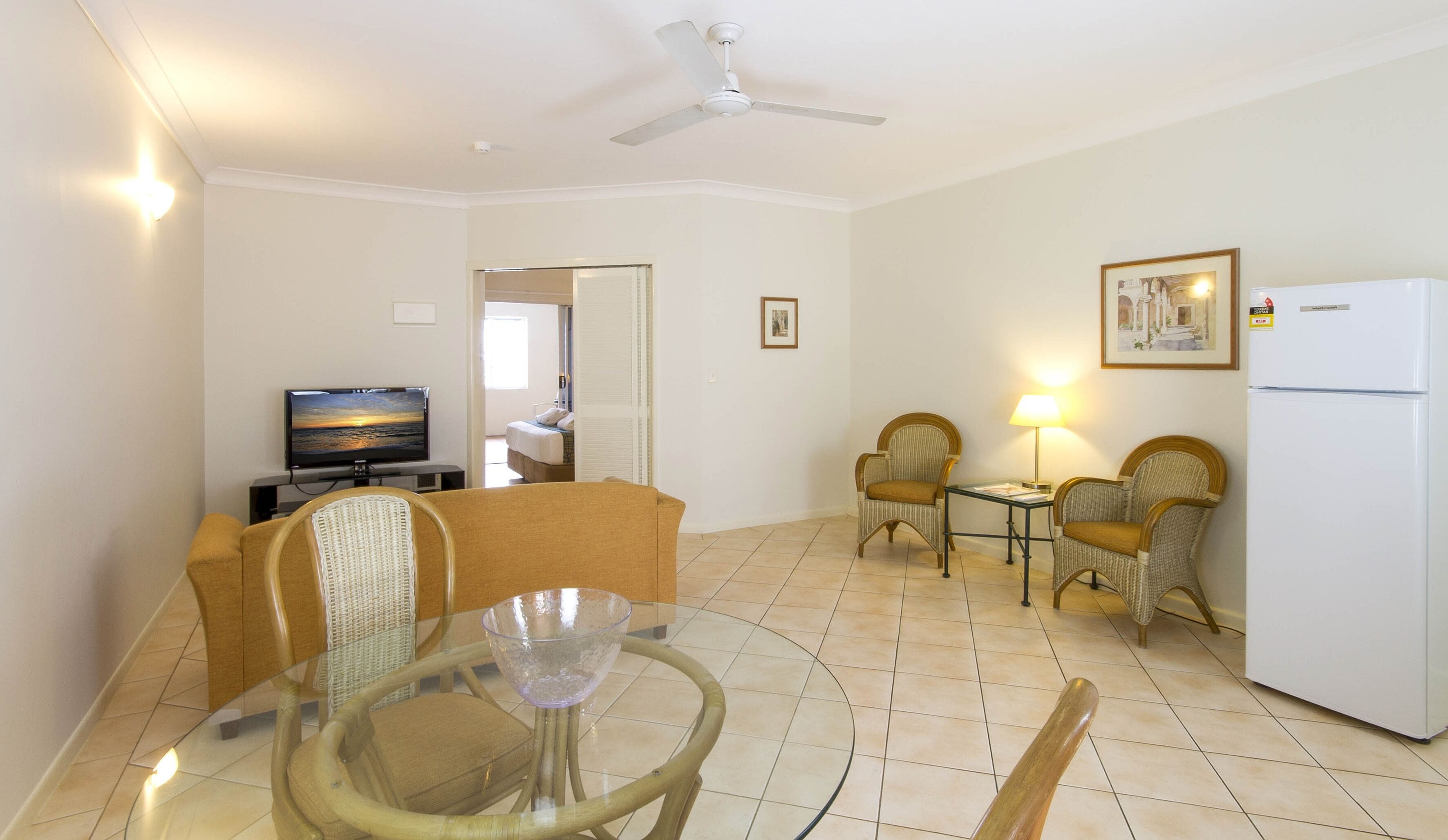 Habitación de hotel con televisión, una mesa de comedor con sillas, refrigerador y ventilador de techo.