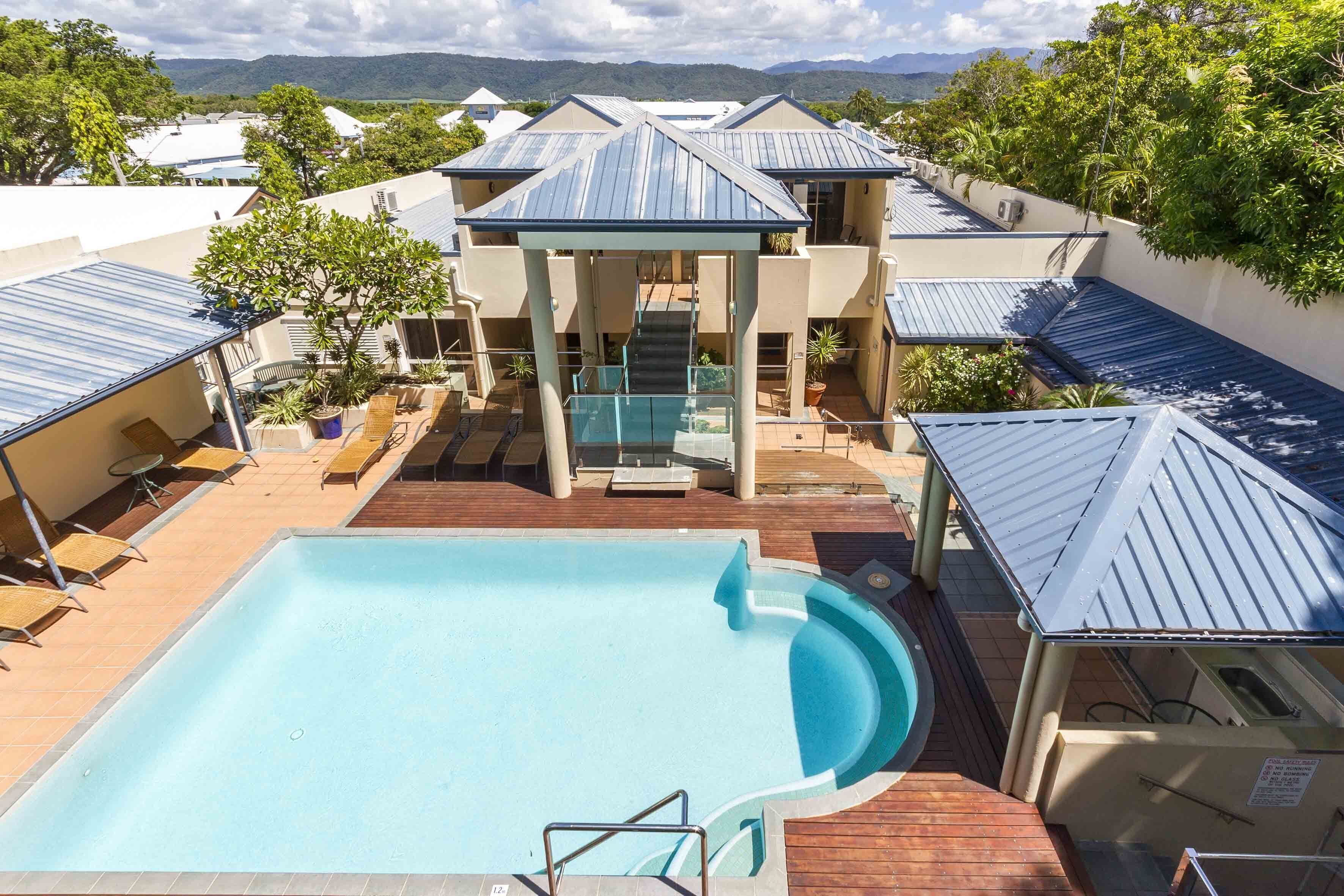 A swimming pool with a wooden deck, a covered seating area, and a view of surrounding buildings and mountains.