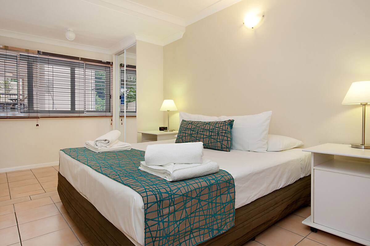 A hotel room with a bed, a nightstand, a lamp, a window with blinds, and a view of greenery outside.