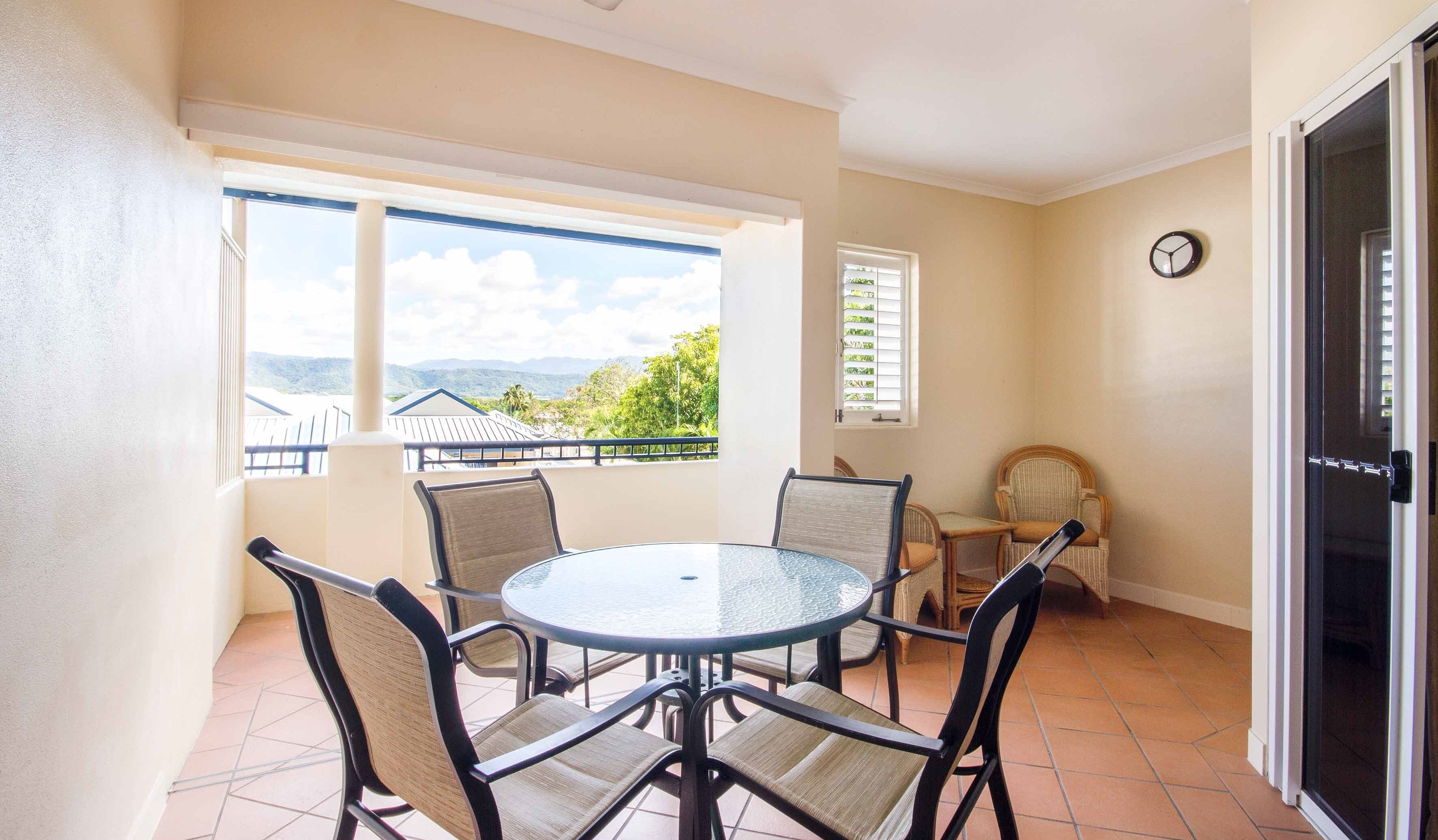 A balcony with a round glass table and four chairs, a window with blinds, a door, and a wall clock.
