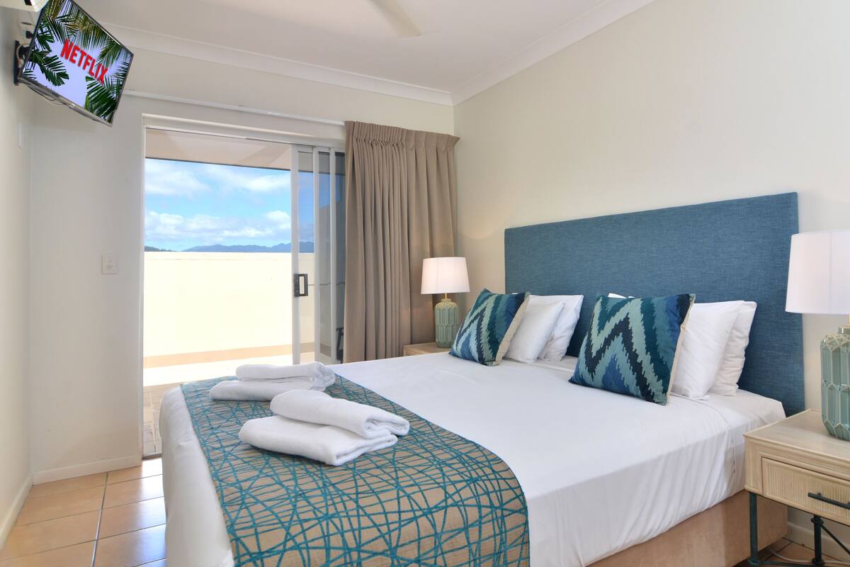 A hotel room with a bed, two bedside tables, a TV, and a view of the outdoors.