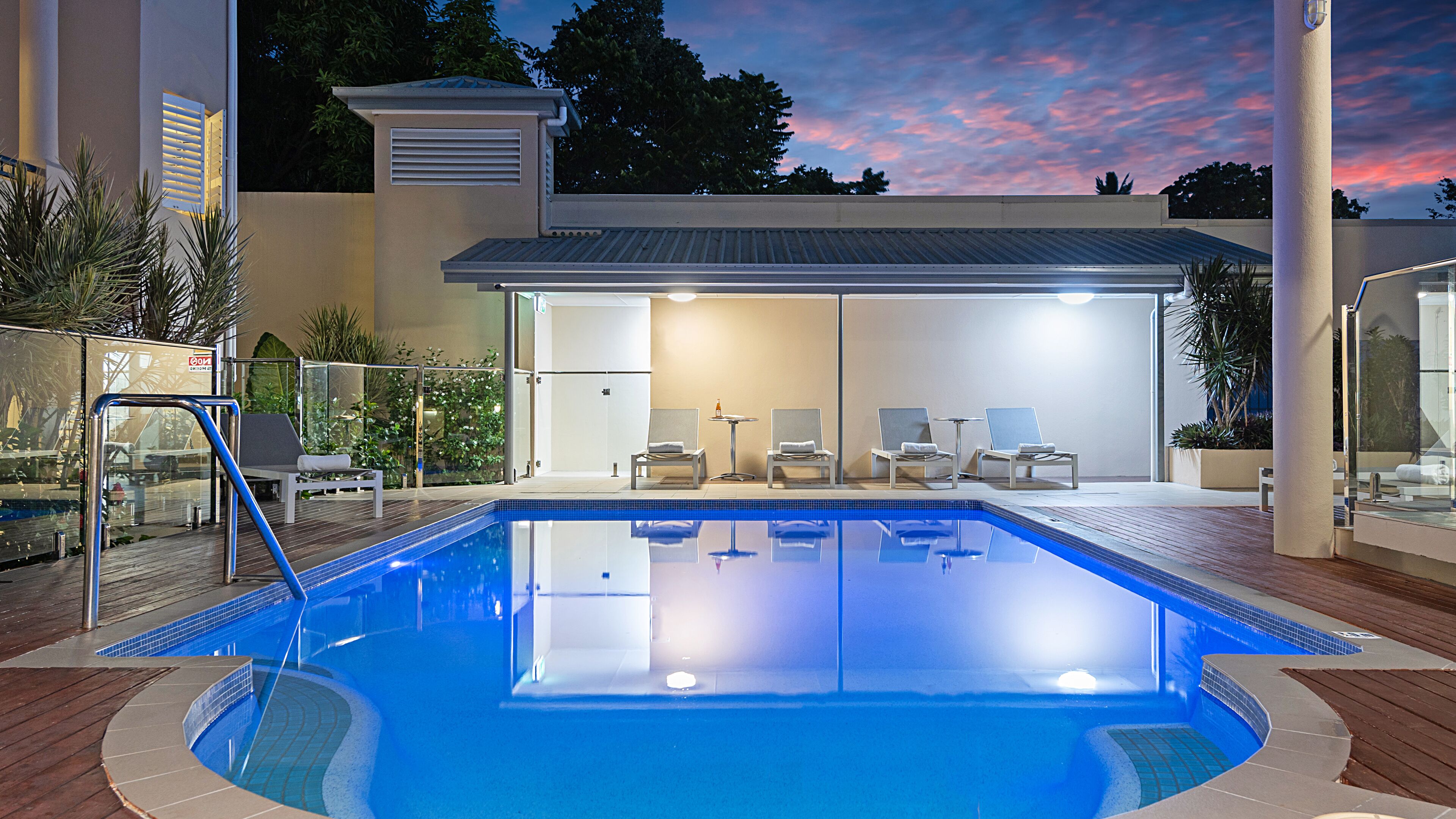 A swimming pool with lounge chairs and a table, surrounded by a deck and plants.