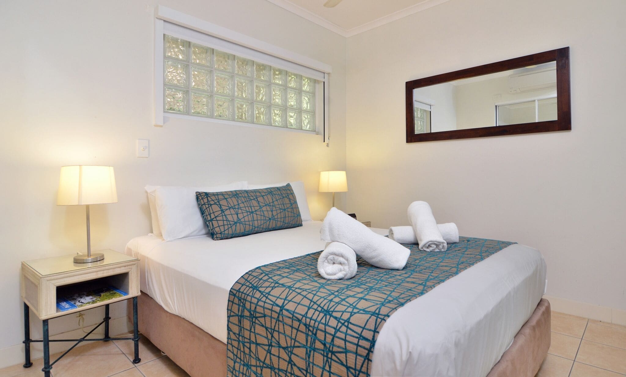 A neatly made bed with a patterned bedspread, two bedside lamps, a small bedside table with a lamp, a window with frosted glass, and a mirror on the wall.