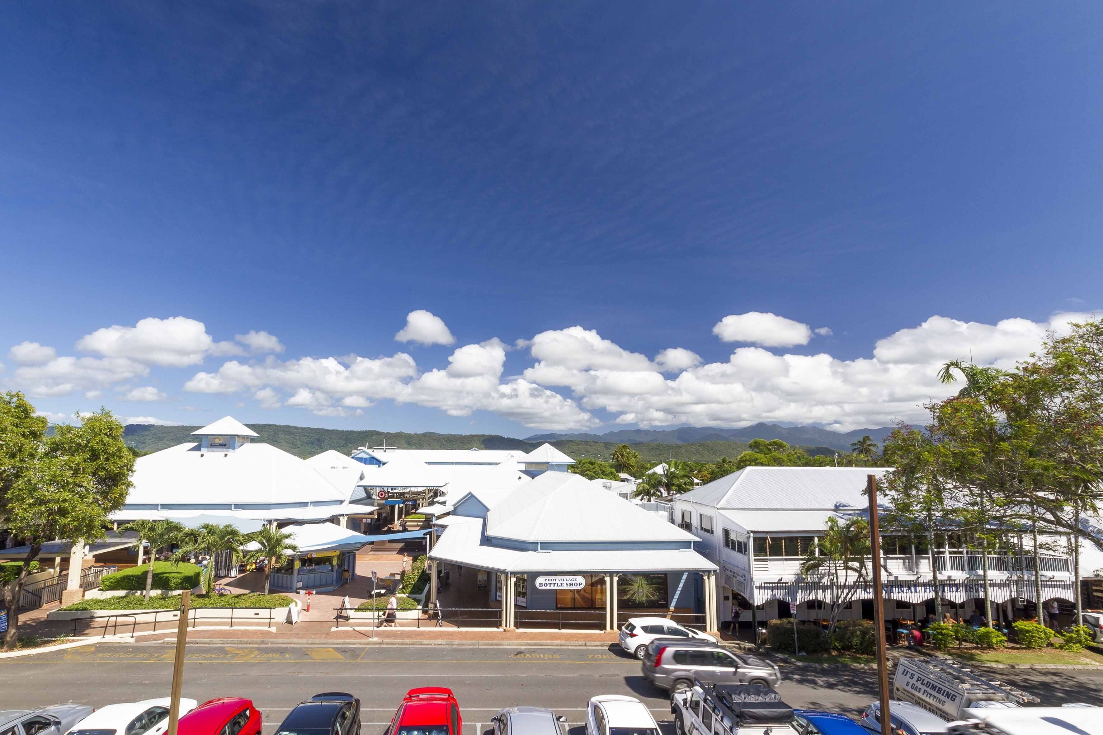 A parking lot with several cars, a building with a sign that reads "It's Plumbing & Gas Fitting," and a mountainous landscape in the background.