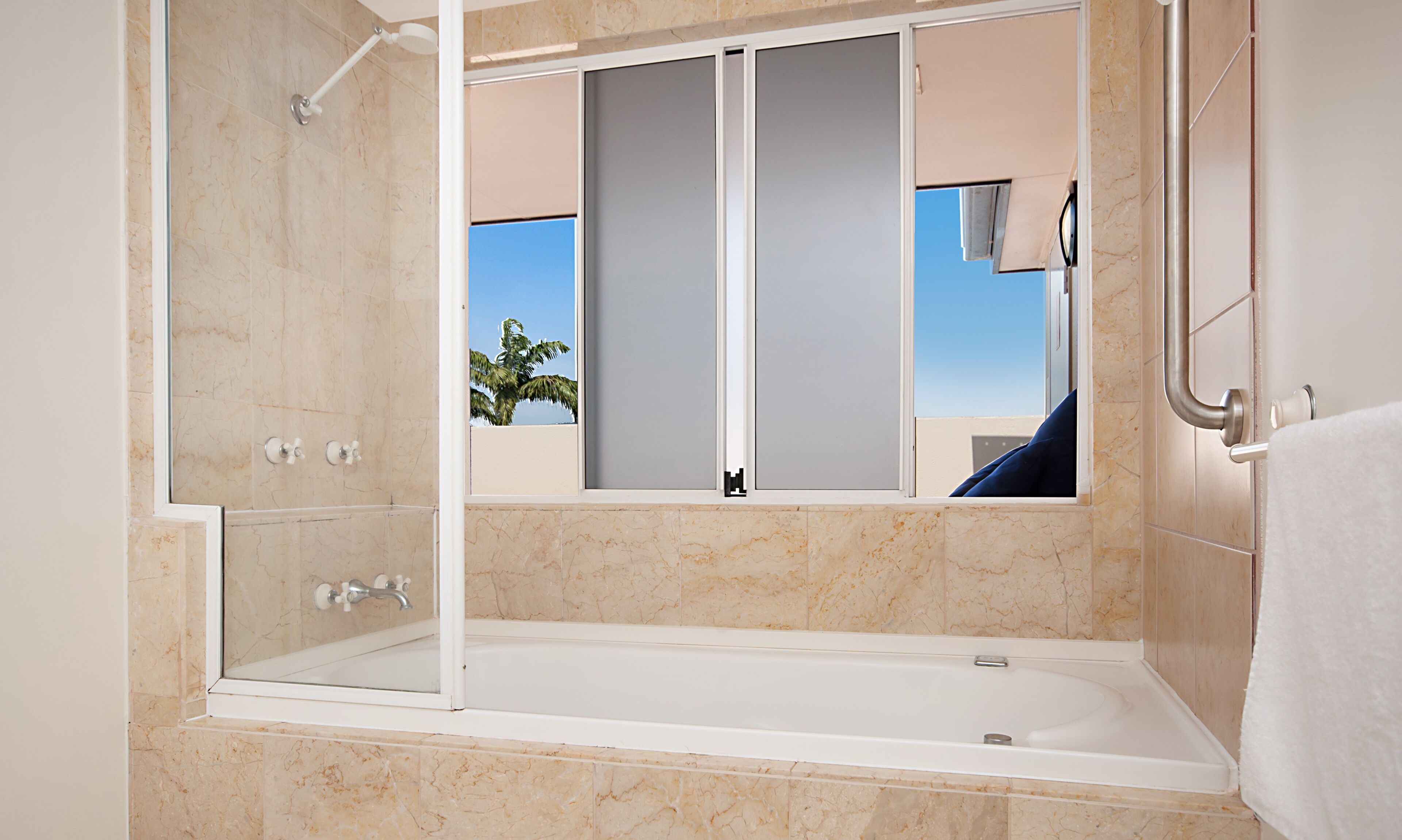 Ein modernes Badezimmer mit einer in Glaswänden gefassten Dusche, einer Badewanne und einem Fenster mit Blick auf Palmen und klaren Himmel.