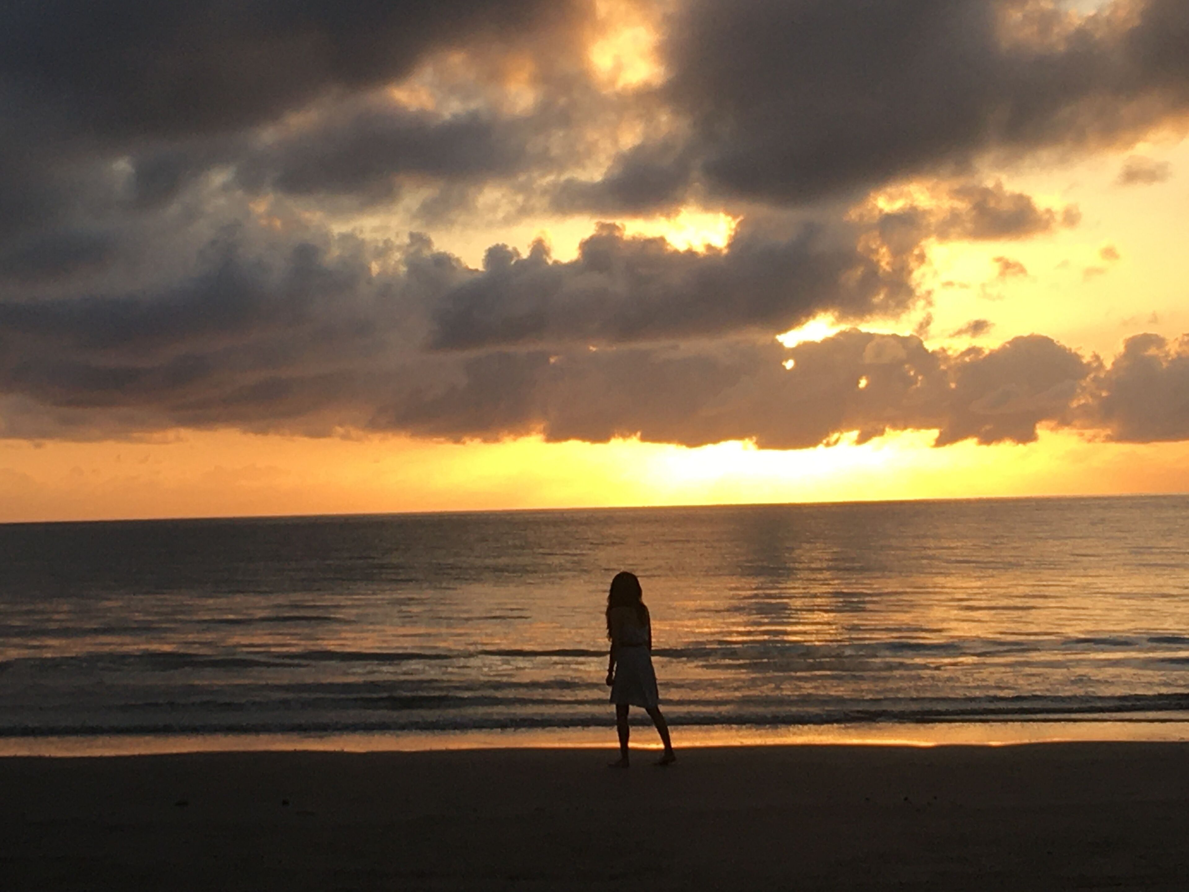 Een zonsondergang aan het strand, met een persoon die langs de waterlijn wandelt.