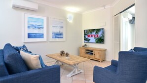 Ein Wohnzimmer mit zwei blauen Sofas, einem Holztisch, einem Fernseher an der Wand und zwei gerahmten Bildern.