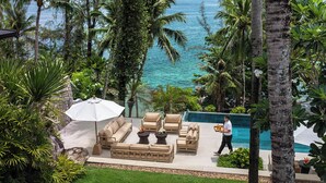Un espace aménagé au bord de la piscine, avec des chaises longues et une table, entouré de palmiers et offrant une vue sur l’océan.