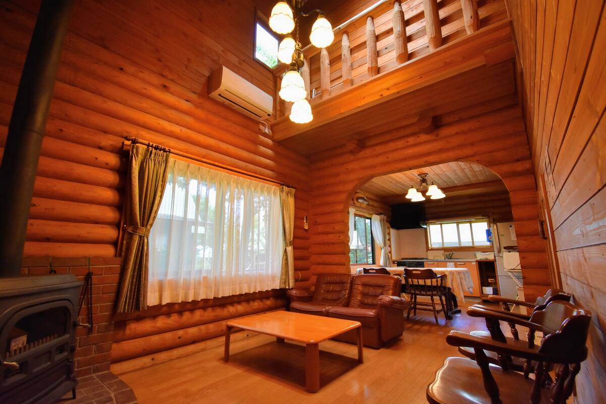 A log cabin interior with a fireplace, wooden furniture, and a kitchen area.