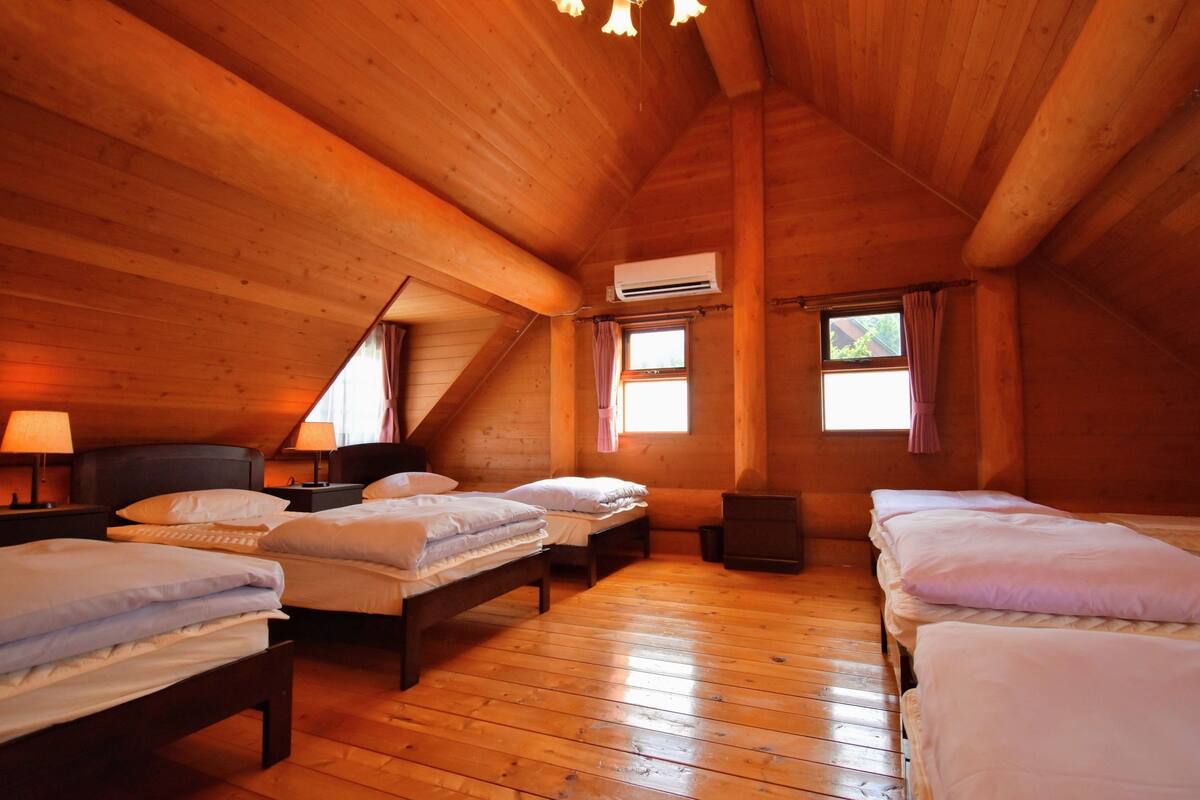 A cozy attic bedroom with wooden walls and floors, two beds, a window with curtains, and an air conditioning unit.