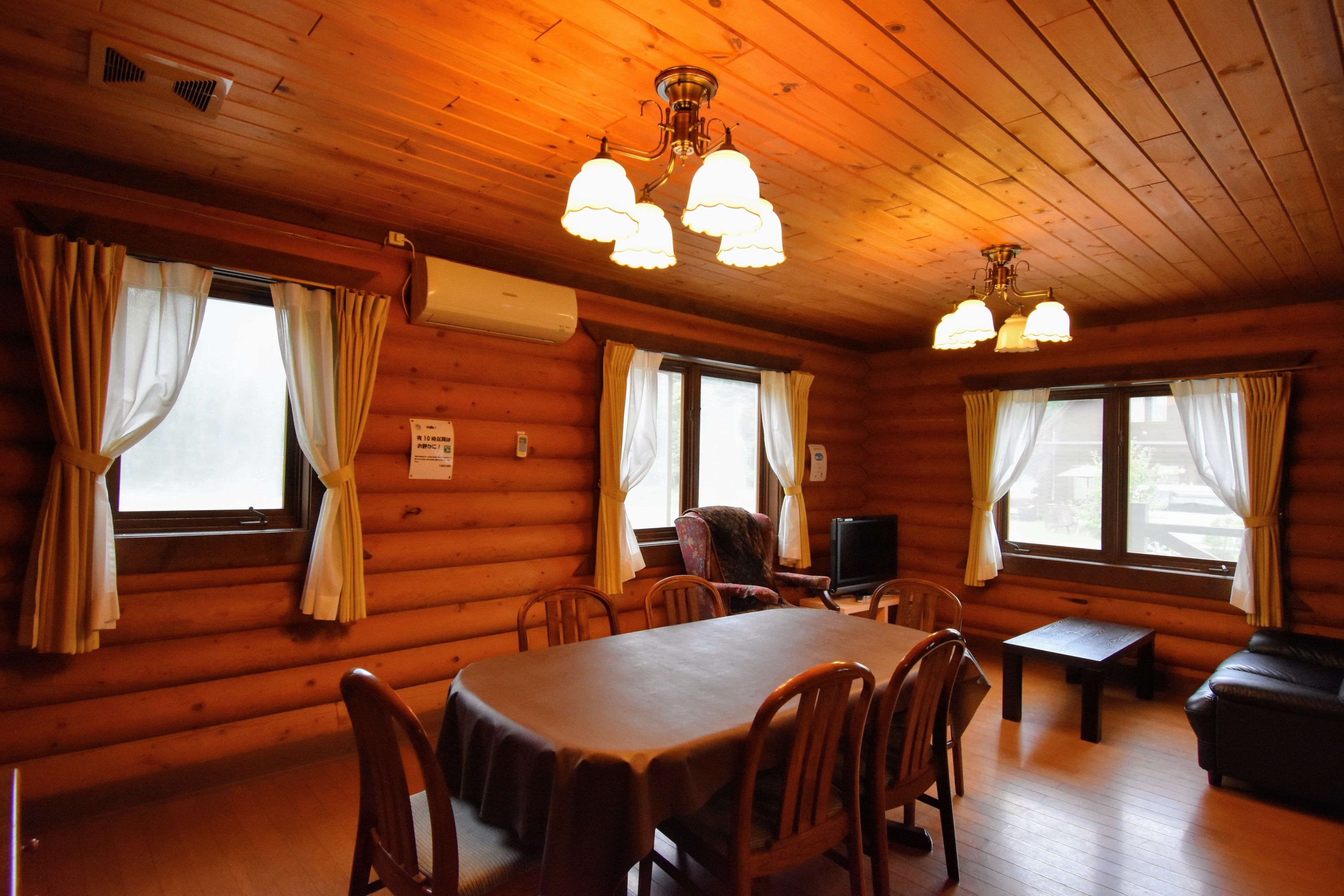 A wooden cabin interior with a dining table, chairs, a sofa, and a television.