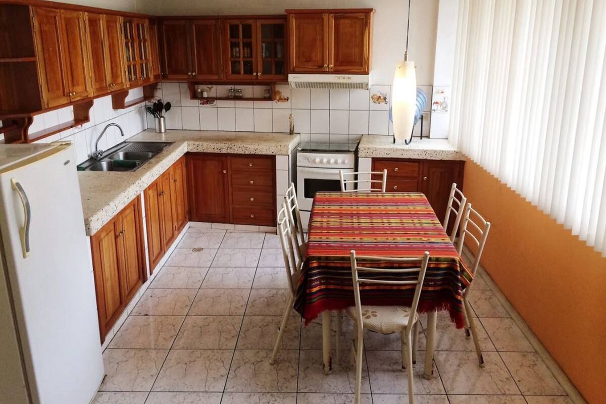 Een kleine keuken met houten kastjes, een spoelbak, kookplaat en een eettafel met stoelen.