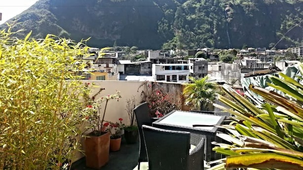 A rooftop terrace with potted plants and outdoor furniture, overlooking a cityscape with buildings and a mountain in the background. - Mayra's Spanish School (Baños de Agua Santa)