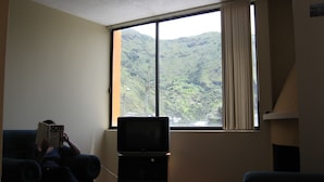 A living room with a television on a stand, a person reading a book, and a window with a view of green hills. - Mayra's Spanish School (Baños de Agua Santa)