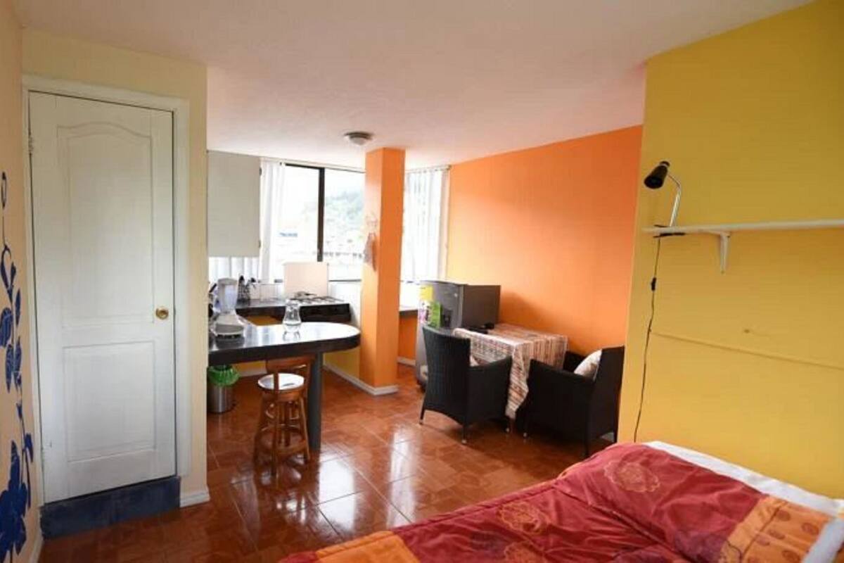 Een knus kamertje met een bed, een klein eettafeltje, een kitchenette en een raam.