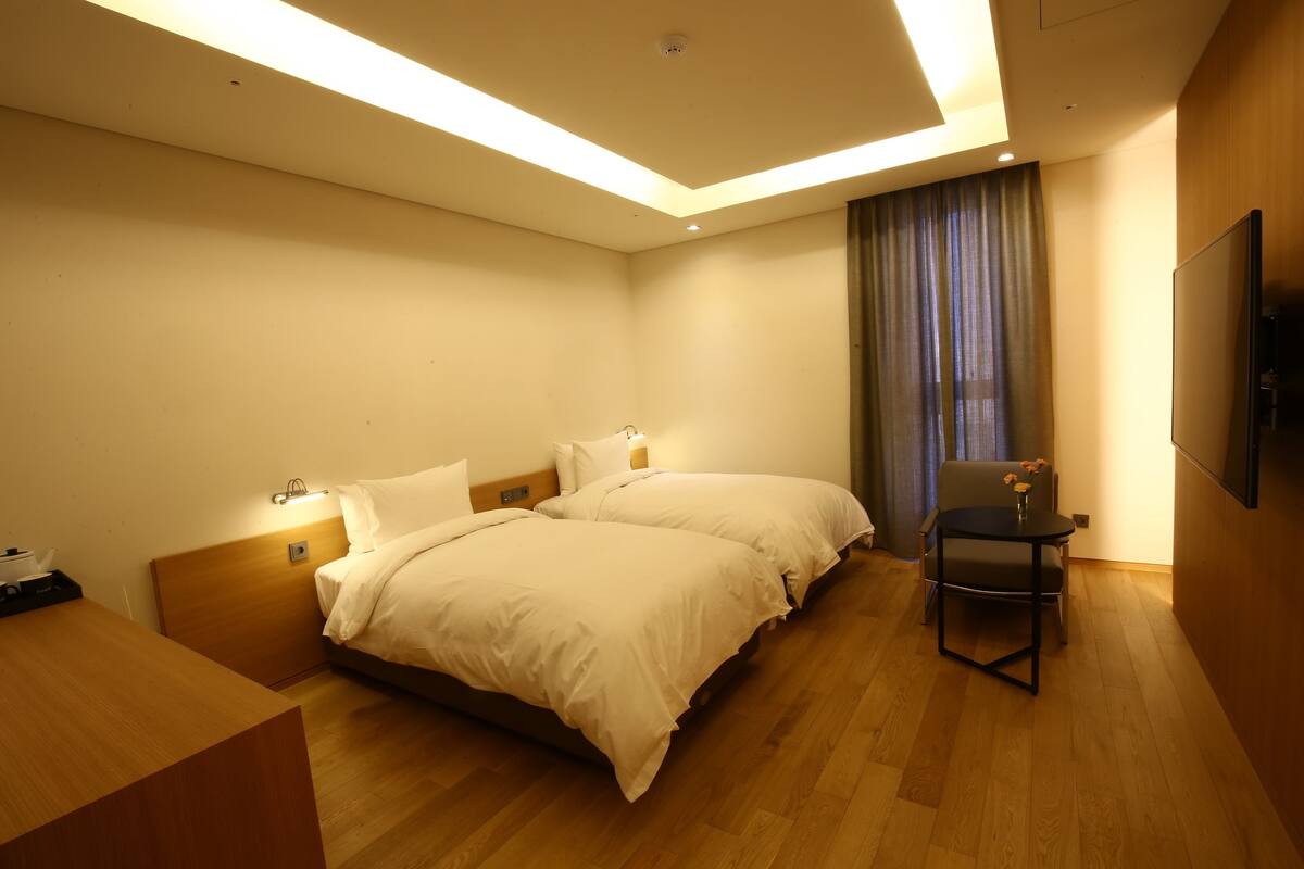 Quarto de hotel com duas camas, uma escrivaninha, uma cadeira, televisão de tela plana e uma janela grande com cortinas.