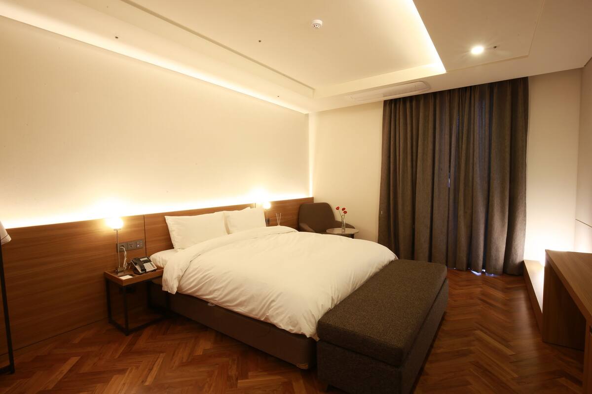 Quarto de hotel com uma cama grande, uma mesinha de cabeceira com luminária, uma pequena mesa com um vaso de flores e um banco de madeira.