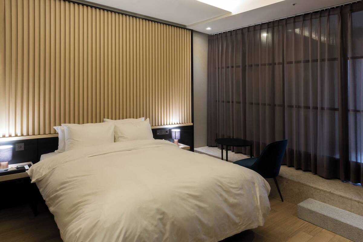 Quarto de hotel com cama grande, mesa de cabeceira, cadeira e janela com cortinas.