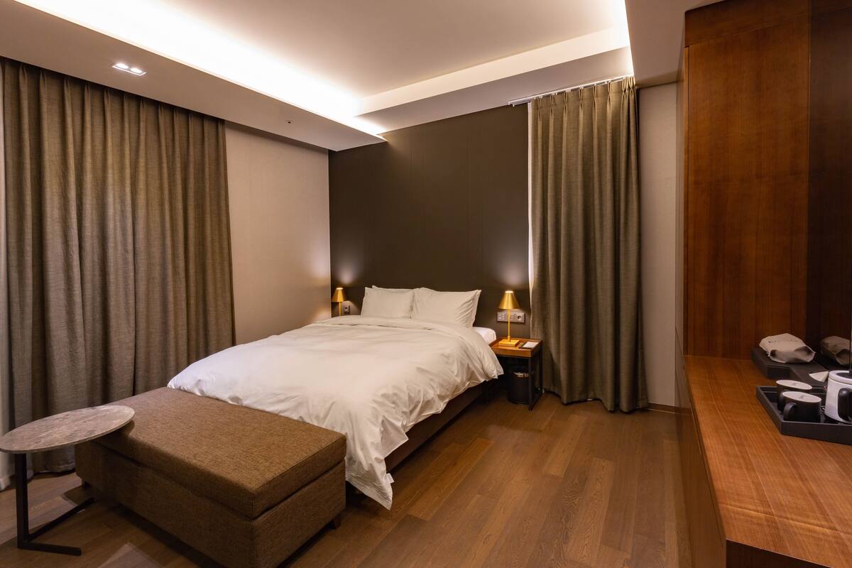 Quarto de hotel moderno com uma cama grande, mesas de cabeceira, uma pequena mesa e um banquinho.