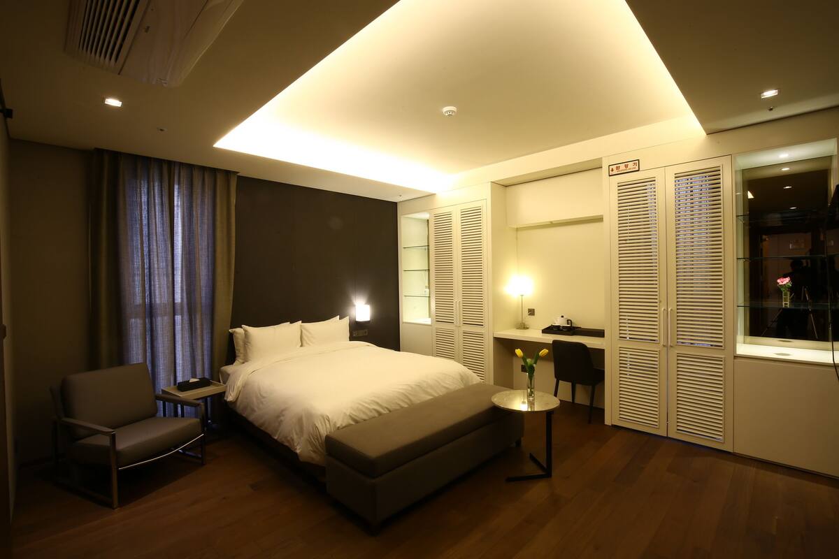 Quarto de hotel moderno com uma cama grande, mesa de cabeceira, cadeira e uma pequena escrivaninha com abajur.
