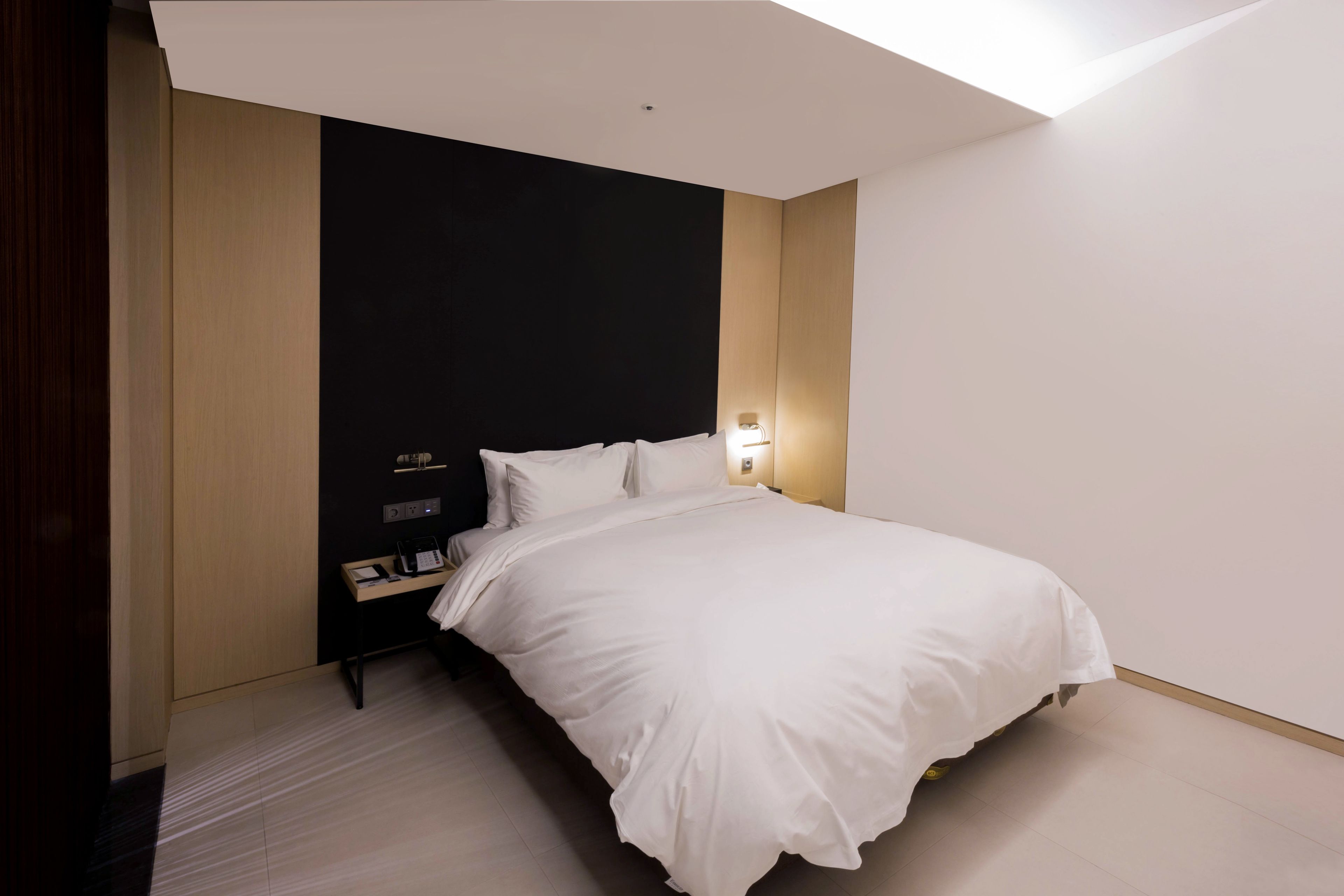 A modern hotel room with a large bed, a bedside table, a nightstand, and a wall-mounted light.