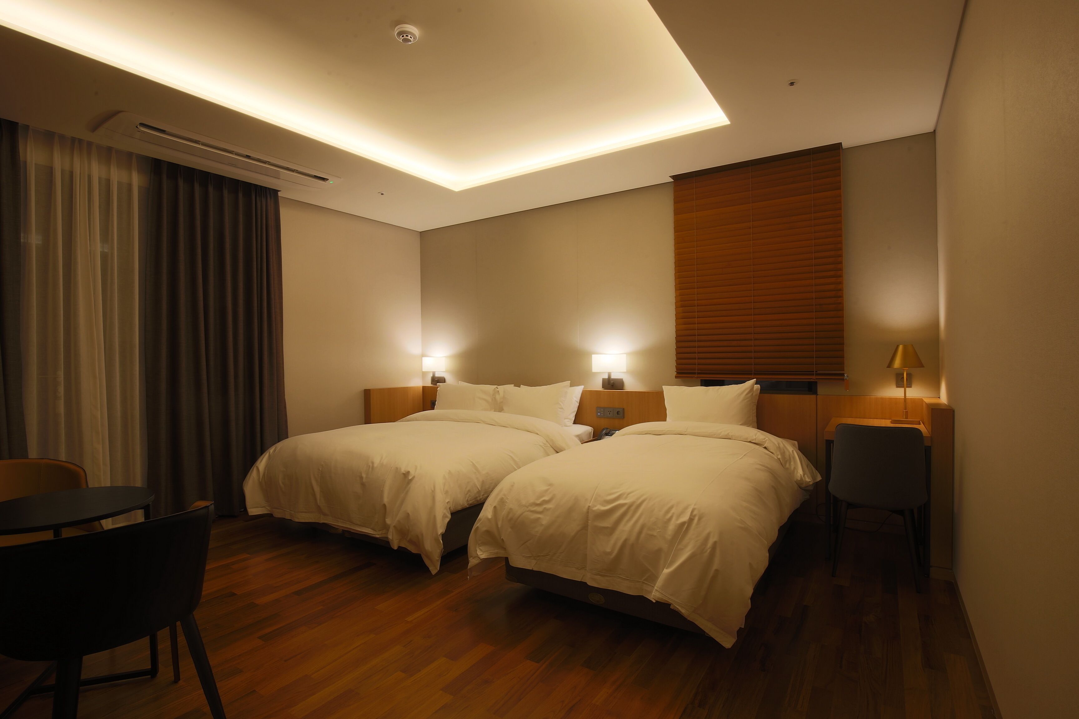A hotel room with two beds, a desk, a chair, and a window with blinds.