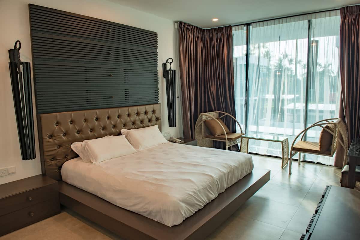 Een moderne hotelkamer met een groot bed, een hoofdbord met horizontale latten, een groot raam met gordijnen en een zithoek met een stoel en een tafeltje.