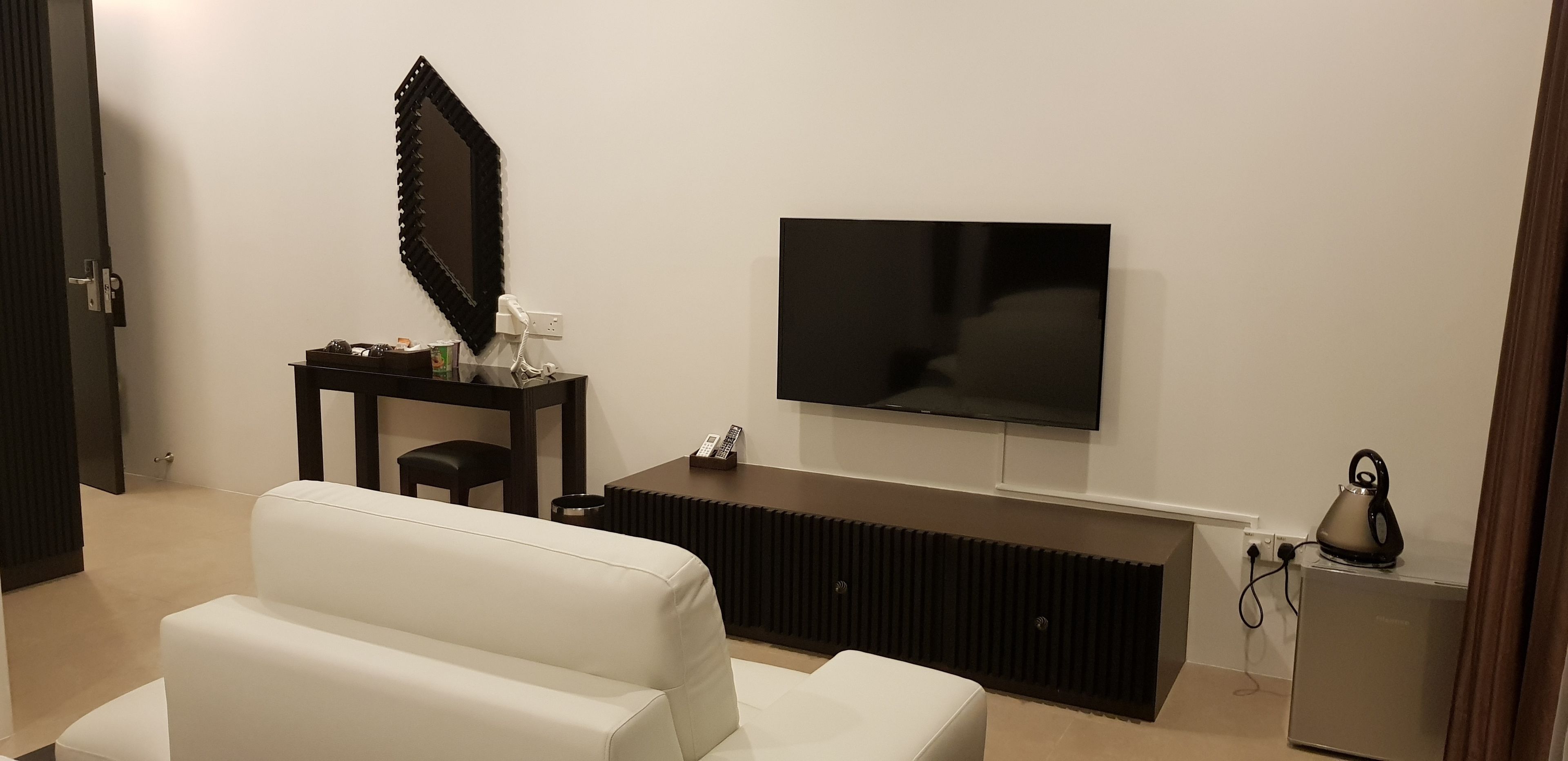 A modern living room with a flat-screen TV mounted on the wall, a white sofa, a dark wooden coffee table, and a small cabinet with a kettle.