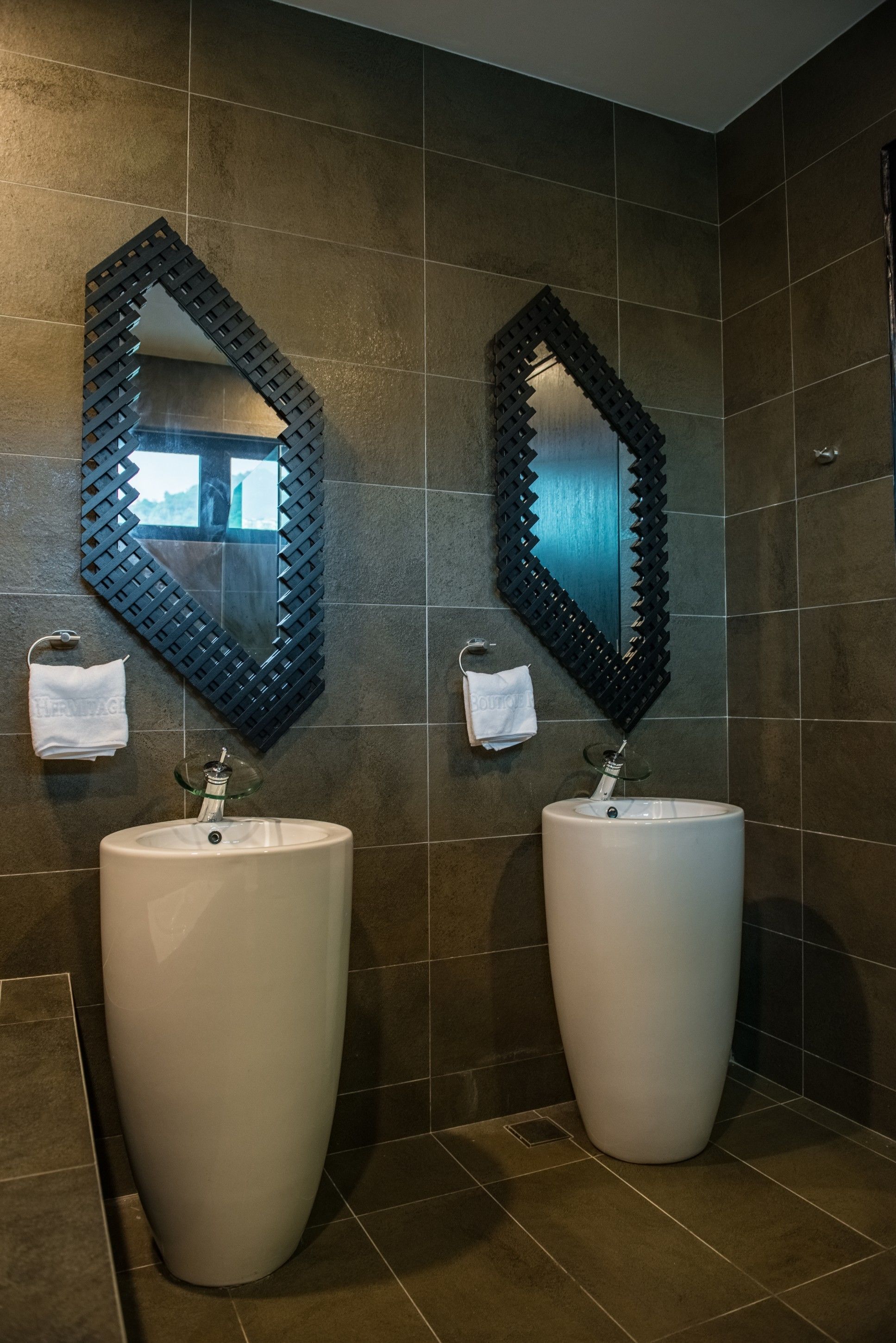 Un baño moderno con dos lavamanos grandes de color blanco, paredes cubiertas de azulejos oscuros y espejos decorativos con marcos negros.
