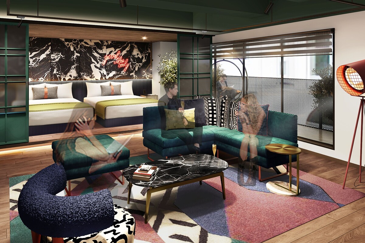 Una sala moderna con sofá, sillón y una mesa de centro, una cama con cabecera verde, y un ventanal con persianas.