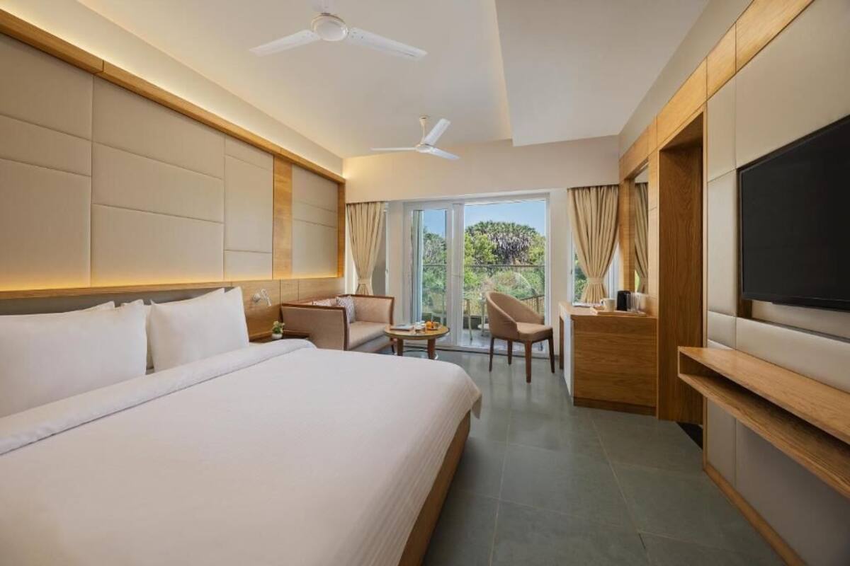 A hotel room with a large bed, a TV, a ceiling fan, a seating area with a table, and a balcony with curtains.