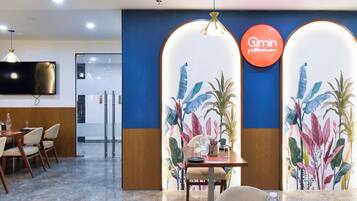 A modern hotel lobby with a dining area, a large screen TV, and a sign reading "Qmin".