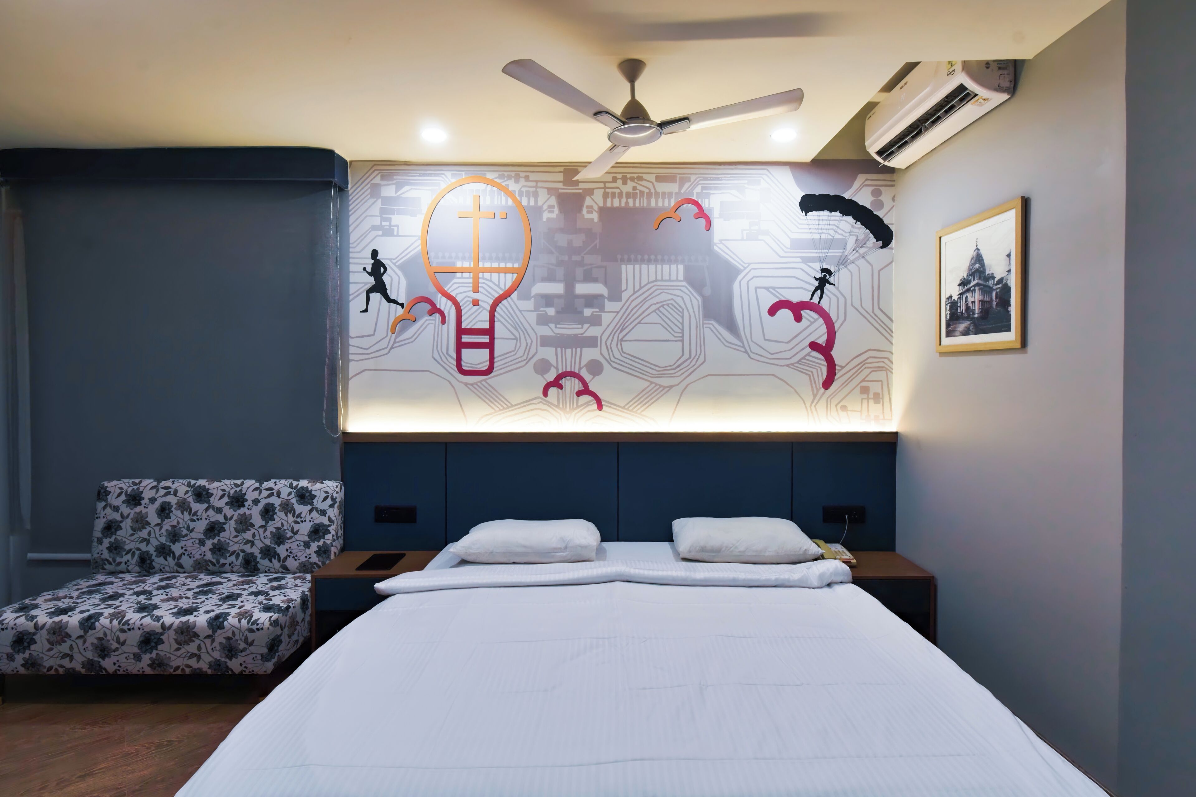 A modern bedroom with a large bed, a ceiling fan, an air conditioning unit, a wall-mounted TV, a framed picture, and a patterned chair.