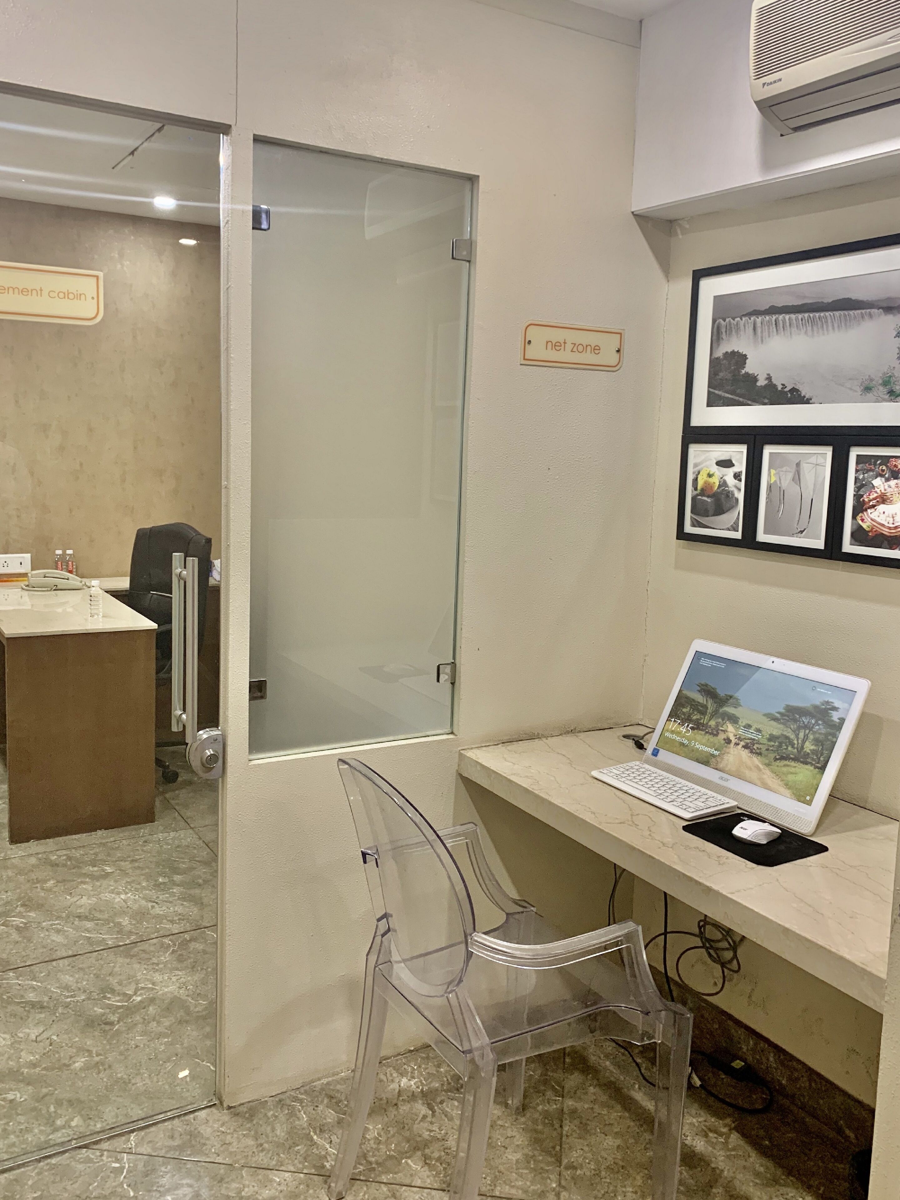 Un espace de bureau moderne avec une cloison en verre, un bureau avec un ordinateur portable, une chaise en plastique transparent et un tableau représentant une cascade accroché au mur.