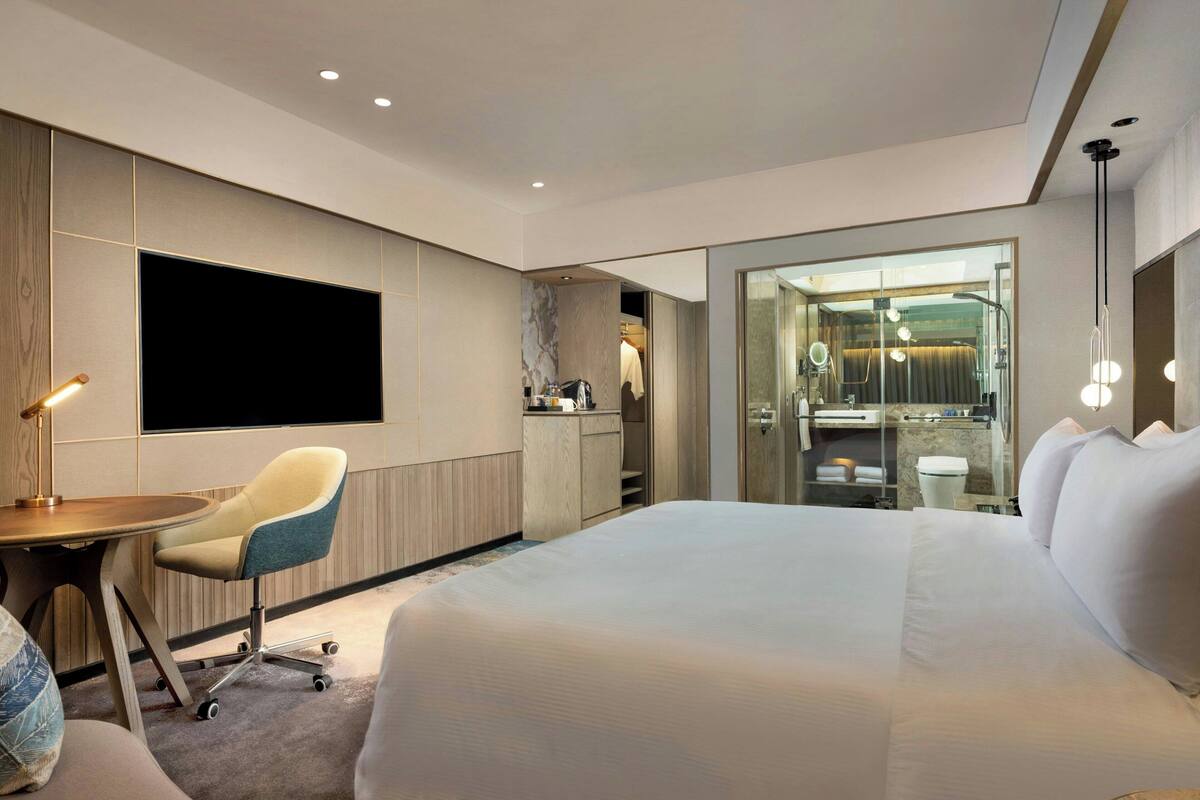 A modern hotel room with a large bed, a desk with a chair, a TV, and a glass-enclosed bathroom.