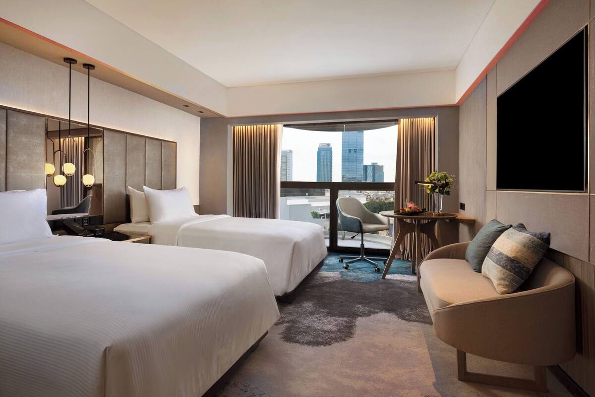 Quarto de hotel com duas camas, uma escrivaninha, uma cadeira, uma janela grande com vista para a cidade e uma TV de tela plana.