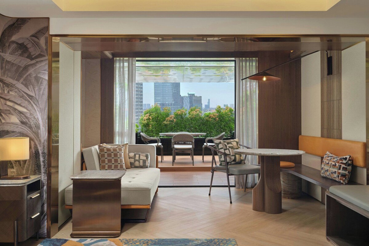 A modern living room with a sofa, a coffee table, and a dining area with chairs. There is a large window with a view of buildings and greenery outside.